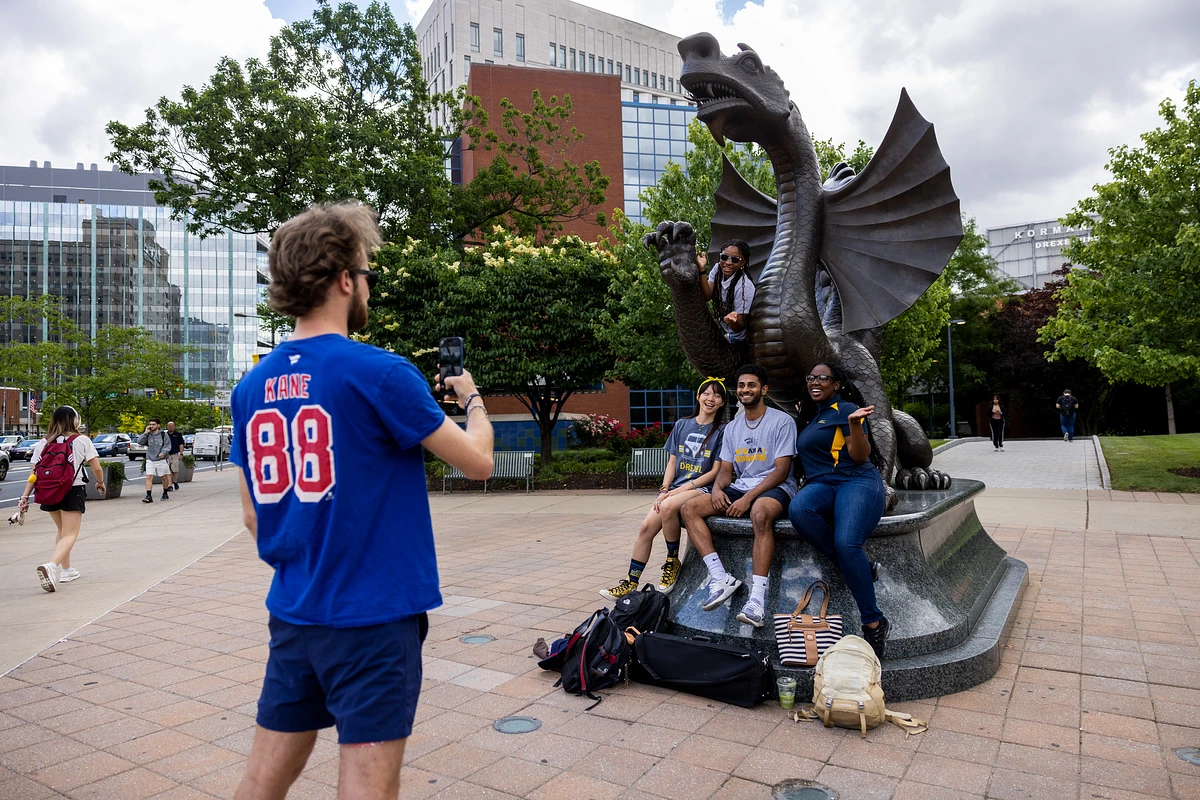 A person is taking a photo of four friends sitting on a dragon statue outside a building.