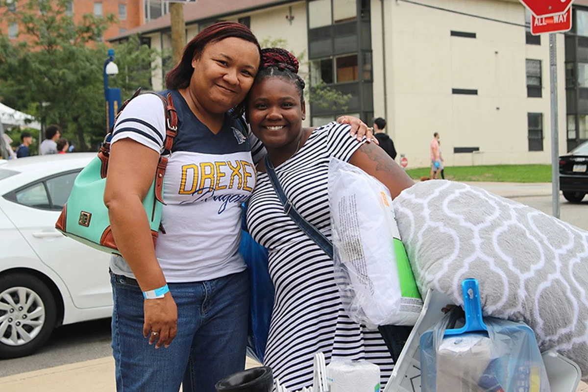 Two smiling people pose together, one carrying bedding and household items, outside a building on a sunny day.