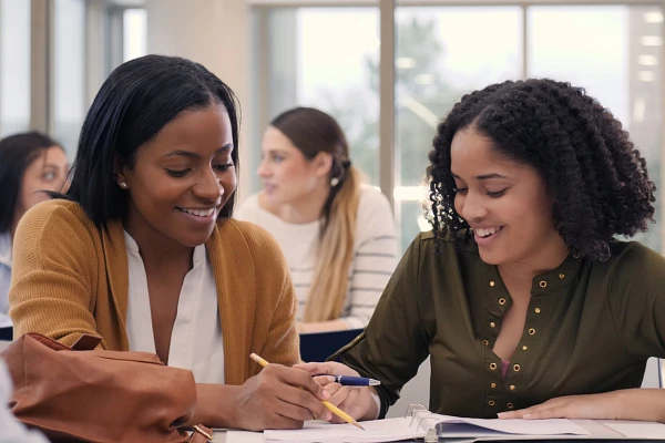 Two students working together with a binder open in front of them, two more students behind them working together