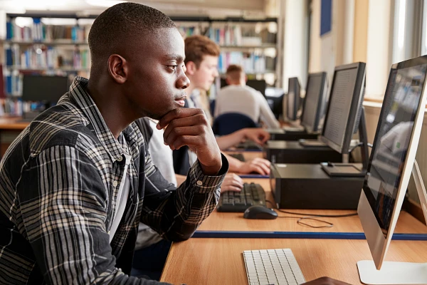 A young man is focused on using a computer in a library or study area with others working in the background.