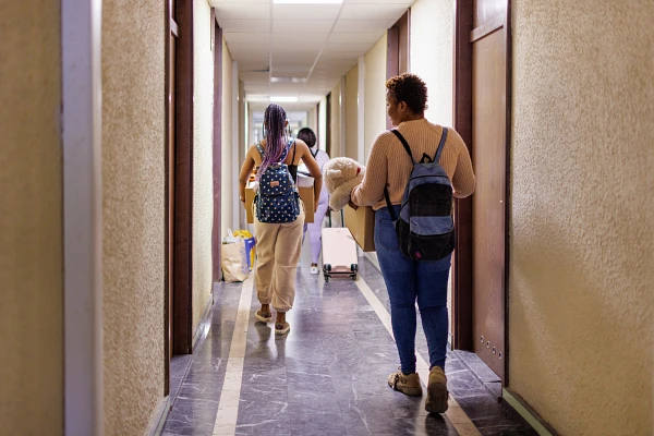 Two people with backpacks walk down a hallway, carrying luggage and a plush toy.