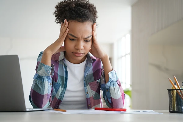 A person sits at a desk, looking stressed while holding their head in their hands.