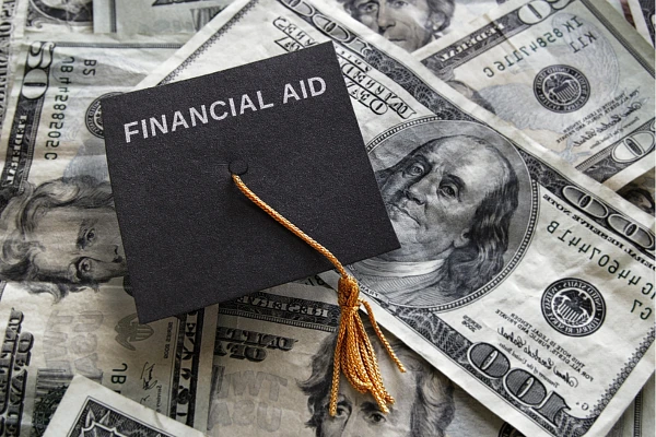 A graduation cap with the words "FINANCIAL AID" rests atop a pile of U.S. dollar bills.