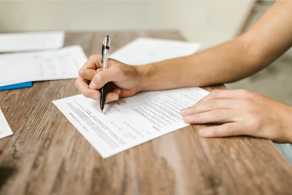 A person is filling out a form on a wooden table using a black pen.
